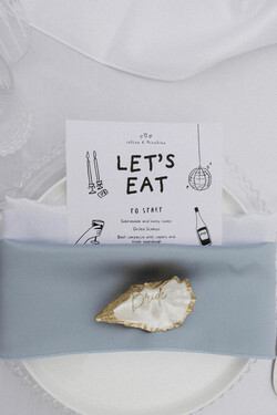Decorative plates with light blue and white napkins on top on a white linen with stationary and oyster shell that that says &quot;Bride&quot;
